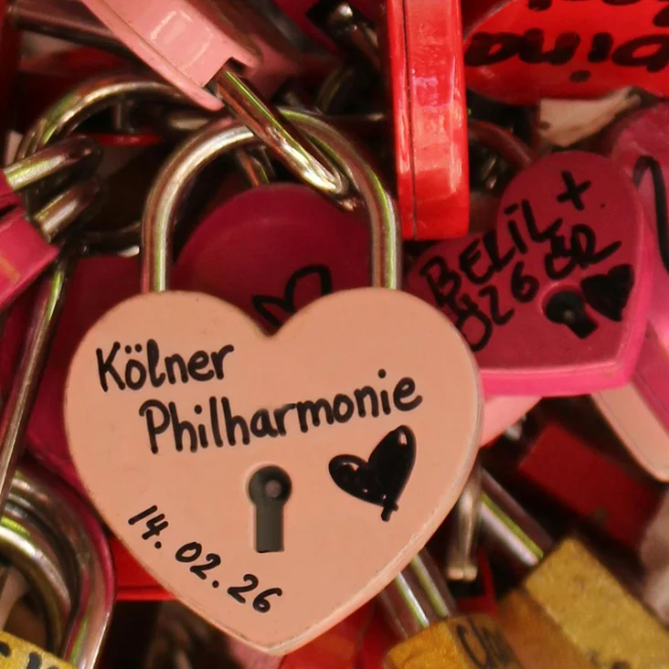 Ein herzförmiges Vorhängeschloss mit der Aufschrift "Kölner Philharmonie". Es ist mit anderen bunten Liebesschlössern an einem Geländer angebracht.