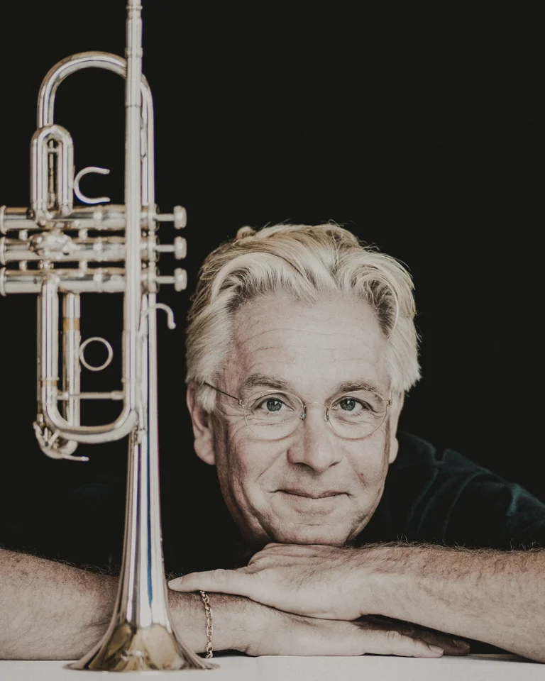 Håkan Hardenberger: A man with gray hair leans on a white pedestal, with a trumpet standing upright beside him.