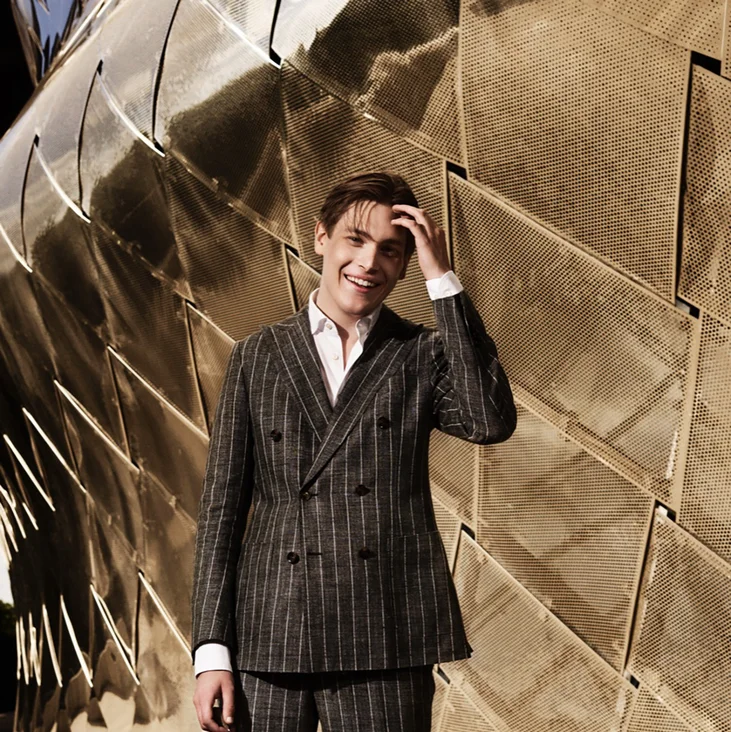 Ein Mann (Klaus Mäkelä) in einem gestreiften Anzug steht vor einer strukturierten, goldenen Wand und lächelt in die Kamera.