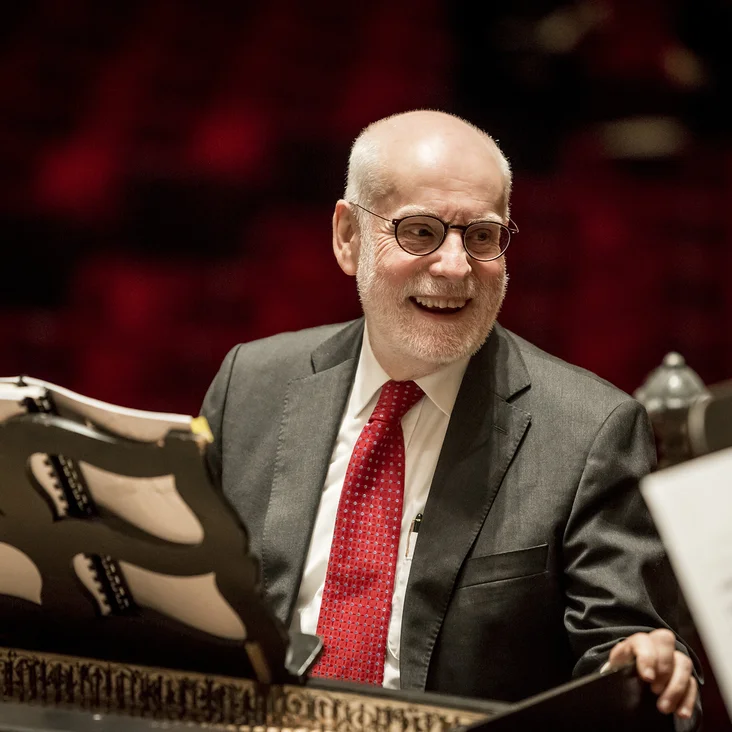 Ton Koopman: Ein Mann in Anzug und roter Krawatte lächelt am Cembalo, mit Notenständer und verschwommenem Hintergrund.