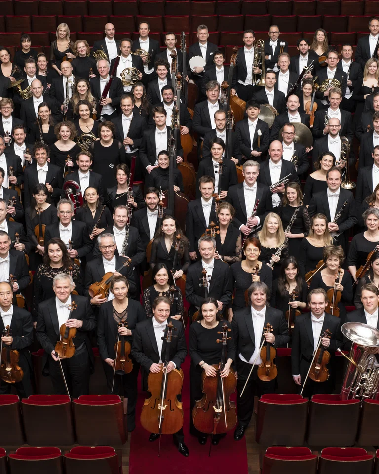Royal Concertgebouw Orchestra: Ein großes Orchester in formeller Kleidung mit verschiedenen Instrumenten, posiert in einem Konzertsaal. Die Musiker stehen in geordneter Formation.