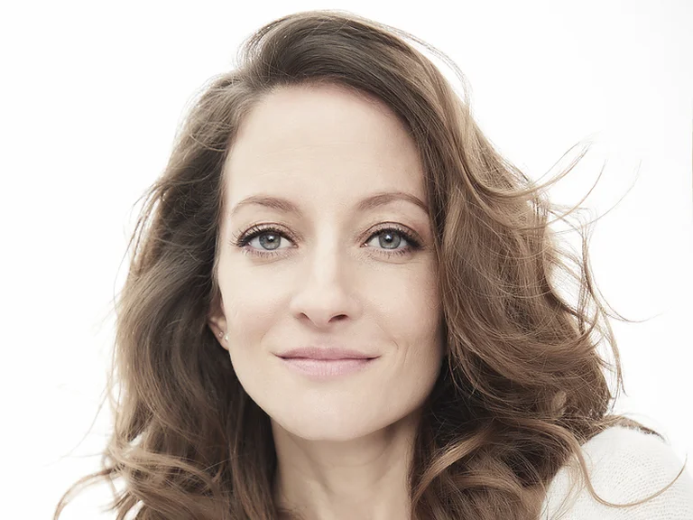 Christiane Karg: Porträt einer Frau mit lockigem Haar und entspanntem Ausdruck in einem weißen Pullover vor hellem Hintergrund.