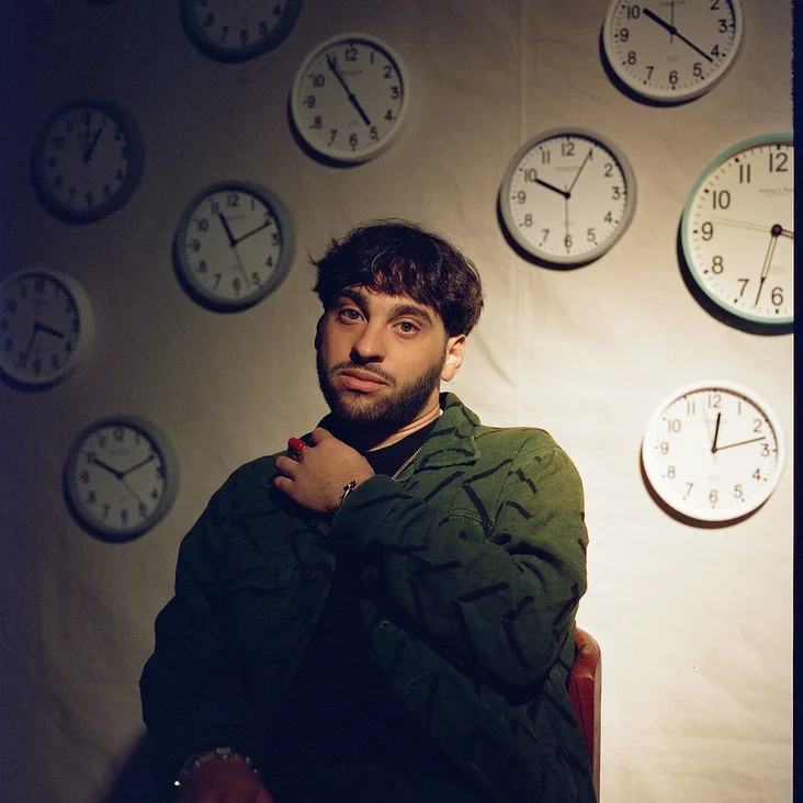 Ein Mann (Samora Pinderhughes) sitzt vor einer Wand mit vielen analogen Uhren. Er trägt einen dunkelgrünen Pullover und hat einen nachdenklichen Ausdruck.