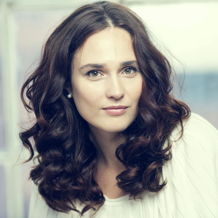 Frau mit braunen, welligen Haaren, trägt ein weißes Oberteil. Sie schaut direkt in die Kamera und lächelt leicht.