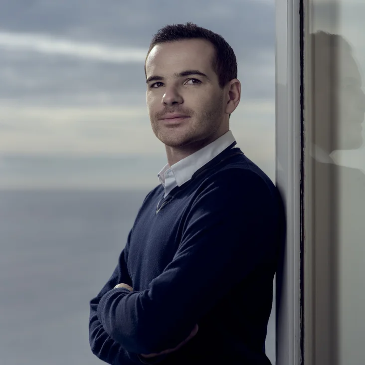 Ein Mann (Lionel Bringuier) steht vor einem Fenster mit Blick aufs Meer. Er trägt einen Pullover und blickt nachdenklich zur Seite.
