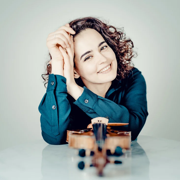 Alena Baeva: Frau mit lockigem Haar lächelt, lehnt sich an einen Tisch mit einer Geige, trägt ein blaues Hemd und wirkt entspannt.