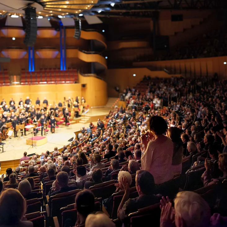Ein Konzertsaal (Kölner Philharmonie) voller Zuschauer applaudiert einer großen Orchesteraufführung. Die Bühne ist hell erleuchtet, die Akustik beeindruckend.