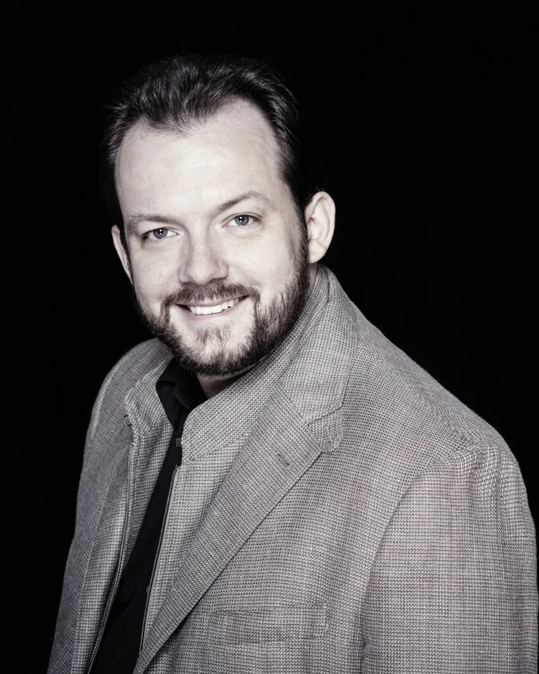Andris Nelsons: Man in a gray suit with a slight smile against a black background. He has a beard and short hair.