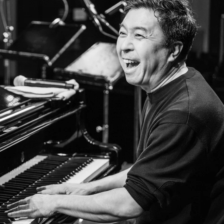 Ein Musiker spielt mit Freude Klavier auf einer Bühne. Noten und ein Mikrofon sind im Hintergrund sichtbar.