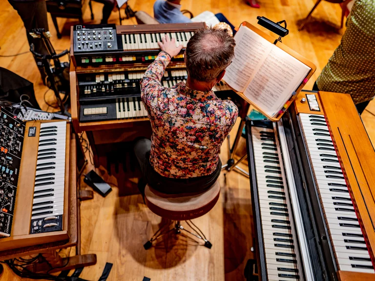 Ensemble Resonanz: Ein Musiker in einem bunten Hemd spielt auf mehreren Keyboard-Instrumenten, umgeben von Notenblättern, in einem Musikstudio.