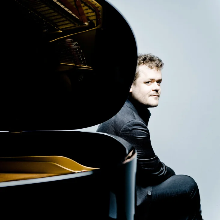 Ein Mann sitzt neben einem Flügel, stilvoll in Schwarz gekleidet. Die Atmosphäre ist modern und elegant.