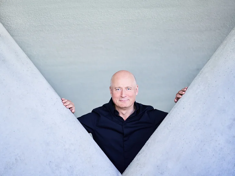 Paavo Järvi: Ein Mann in schwarzer Kleidung steht zwischen zwei großen, grauen Wänden, die ein V formen. Er blickt direkt in die Kamera.