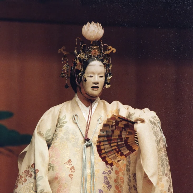 Ein traditioneller Noh-Schauspieler in aufwendigem Kostüm (Hagoromo) und Maske, hält einen Fächer. Der Hintergrund zeigt eine schlichte Bühne.
