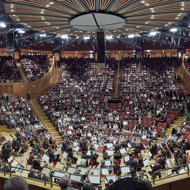 A full concert hall with balconies and a large audience. An orchestra is performing in the foreground.